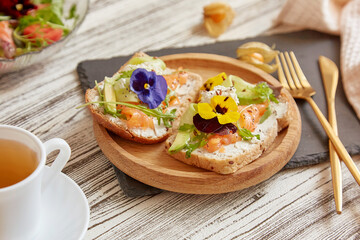 Healthy pescetarian breakfast - toasts with smoked salmon, cottage cheese, edible flowers, avocado and greens. Tea cup. Atmospheric autumn food
