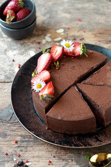 Sliced Chocolate ganache cake with fresh strawberries on a rustic wooden table with red pepper, fresh daisies and black cake stand