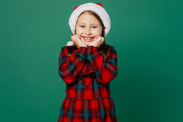 Merry smiling happy satisfied little child kid girl 6-7 years old wear red dress Christmas hat posing look camera prop up hold face isolated on plain dark green background Happy New Year 2023 concept