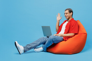 Full body young IT man of African American ethnicity wear red shirt sit in bag chair hold use work...