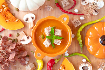Warm and tasty pumpkin soup with croutons and mint on top in pumpkin shaped bowl with spoon surrounded by green and red peppers, mushrooms, garlics, pumpkin seeds on wooden table. High quality photo