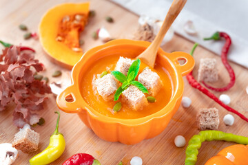 Warm and healthy pumpkin soup with croutons and mint in pumpkin shaped bowl with spoon, red pepper, garlic