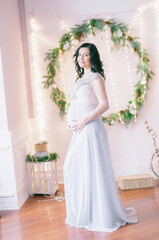 Fototapeta premium Young pregnant woman with dark hair in airy dress in a room decorated with pine needles and sparkling garlands for Christmas. Christmas mood . Pregnancy