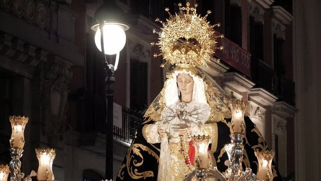 Front view, close-up of Virgin Mary figure at night during a Holy Week procession celebration in Madrid, Spain. Solemn and mournful religious image. Nice city building behind.