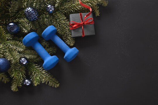 Christmas sport composition with two blue sports dumbbells, gift, fir tree branches and gift on black background. Top view with copy space. Fitness, sport and healthy concept. Christmas greeting card.