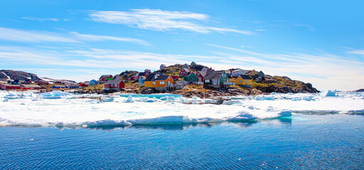 Panoramic view of colorful Kulusuk village in East Greenland - Kulusuk, Greenland - Melting of a iceberg and pouring water into the sea - Drummer statue in the foreground - obrazy, fototapety, plakaty