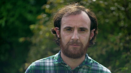 Fototapeta premium Portrait of a casual serious man looking at camera standing outdoors. Redhead 30s male person face closeup with beard