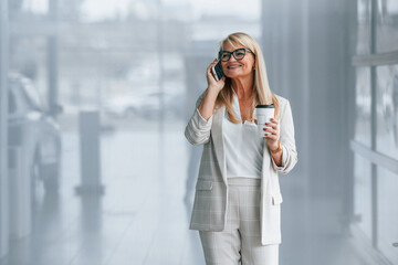 Standing and talking by phone. Woman in white formal clothes is standing indoors