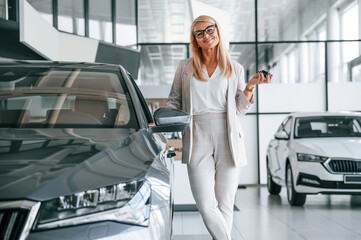 Happy owner of new automobile with keys in hand. Woman in white formal clothes is in the car dealership