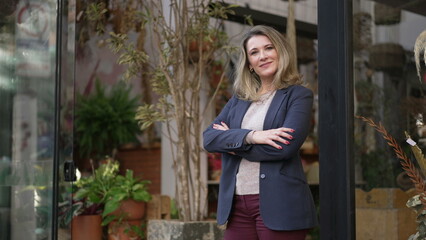Confident mature female entrepreneur standing in front of local small business wearing blue jacket. Happy owner of flower shop standing by sidewalk looking at camera