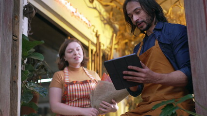 Two young entrepreneur of flower shop. Man calling female work colleague showing tablet device. Employees of small business discussing online orders