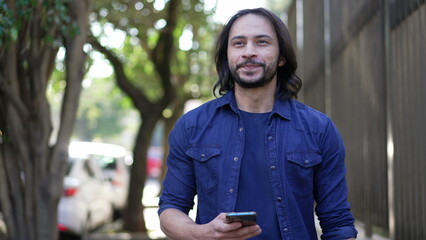 Casual Joyful South American man checking smartphone walking in city street in daylight. Smiling hispanic person using phone