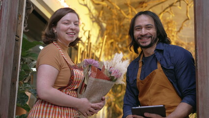 Two employees wearing apron holding tablet smiling at camera. Portrait of workers employees holding flower bouquet arrangement
