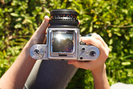 A photographer holding a vintage analog photo camera focusing ajusting taking pictures outside looking through a large square format viewfinder mechanical settings solid metal cogs gagarden background