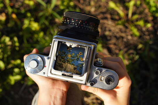 A photographer holding a vintage analog photo camera focusing ajusting taking pictures outside looking through a large square format viewfinder mechanical settings solid metal cogs gagarden background