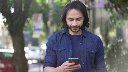 One young hispanic man looking at cellphone device while walking in city street in daylight. South American Brazilian person holding smartphone walks forward in motion