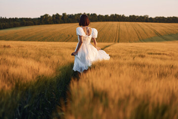 Fototapeta na wymiar Rear view. Beautiful young bride in white dress is on the agricultural field at sunny day