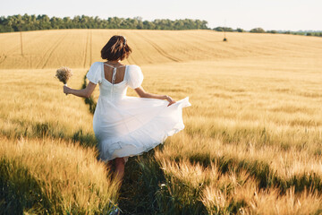 Holding flowers. Beautiful young bride in white dress is on the agricultural field at sunny day