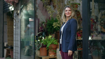 MIddle age entrepreneur smiling standing in the door of her flower shop proud to be the owner of a small business standing by street sidewalk