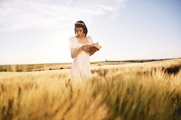 With book in hands. Beautiful young bride in white dress is on the agricultural field at sunny day