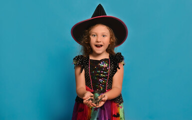 Surprised happy little girl wearing Halloween witch costume holding toy spider and looking at...