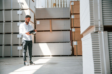 Holding tablet and checking the data. Storage worker is in the warehouse with bunch of products