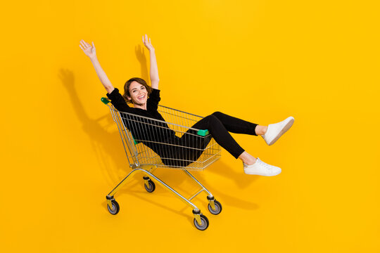 Photo of impressed funny lady wear black shirt rising arms riding shooping tray isolated yellow color background