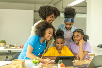 A group of young African kids with their sister use a tablet to communicate with friends. Happy kids use computer for online class under support from a young female teacher. Technology for education