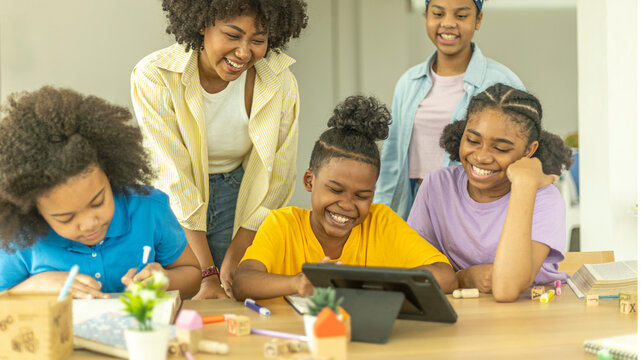 A group of young African kids with their sister use a tablet to communicate with friends. Happy kids use computer for online class under support from a young female teacher. Technology for education