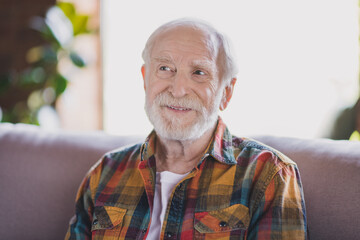 Photo of cheerful positive retired man wear plaid shirt smiling having rest cozy couch indoors apartment