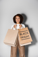 positive young african american woman holding shopping bags with black friday lettering on grey.