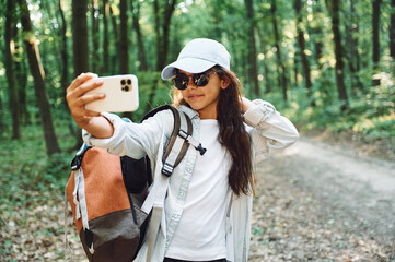 Fototapeta na wymiar Holding smartphone and making selfie. Girl is in the forest at summer day time discovering new places