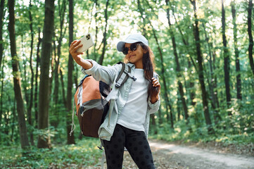 Holding smartphone and making selfie. Girl is in the forest at summer day time discovering new places