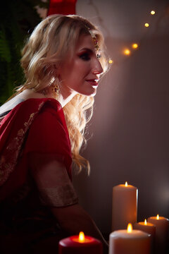 A beautiful European girl looking like an Arab woman in a red room in a harem with candles. The wife creates comfort in the house. Photo shoot of an oriental-style odalisque. A model poses in a sari