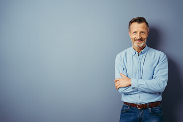 Handsome middle-aged man, standing with his arms folded, looking at camera and smiling. Half-length front portrait against blue background with copy space