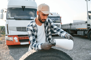 Leaning on the tire, reading the documents. Young truck driver is with his vehicle at daytime