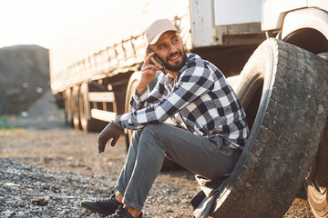 Sitting with phone, waiting for help. Damaged tire. Young truck driver is with his vehicle at...