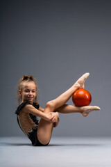 A young girl gymnast in a gymnastic leotard does exercises with a ball. Isolated on a gray...