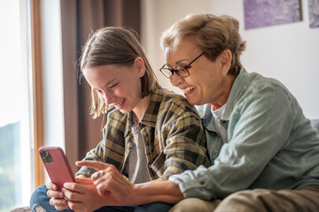 Smiling girl granddaughter with happy grandmother having fun and using looking at smartphone screen...