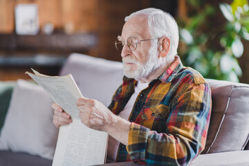 Photo of serious confident retired man dressed plaid shirt spectacles sitting couch reading fresh...