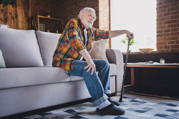 Photo of stressed unhappy man pensioner wear checkered shirt holding walking cane trying standing up sofa indoors house room