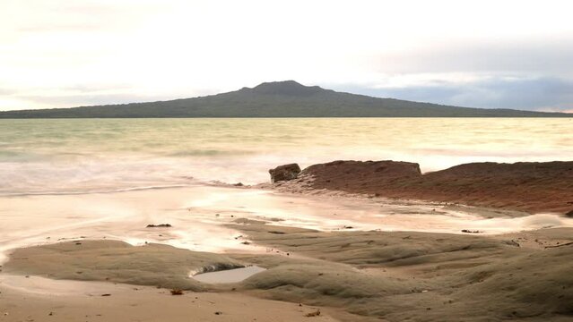 Time lapse of the sun rising behind Rangitoto volcano, viewed from a rocky beach across the turquoise ocean