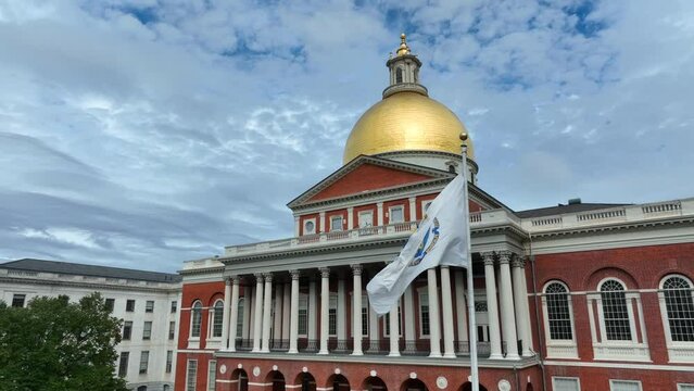 Beautiful orbiting aerial view of Massachusetts state flag flying proudly in front of state house building in Boston, MA. Bright day with blue sky shines on reflective gold dome. Government building.