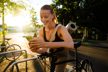 White young woman using cellphone and standing by bicycle after workout
