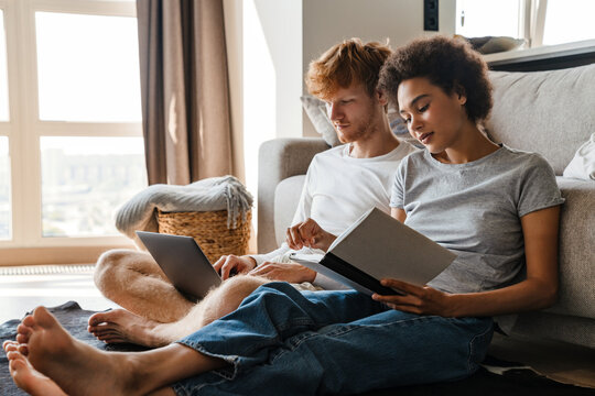 Young multinational couple using laptop and reading book at home