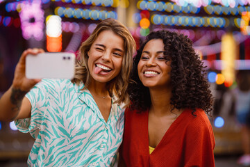Fototapeta premium Young multiracial women taking selfie on cellphone in attraction park