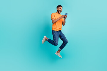 Fototapeta na wymiar Full length photo of cheerful excited man dressed orange t-shirt writing modern device jumping high isolated turquoise color background
