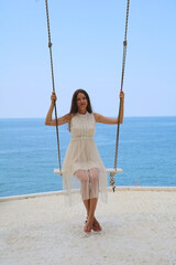 Luxury fashion. Elegant fashion model. Stylish female model. Beautiful girl in a beige dress rides on a swing on the beach against the background of the sea