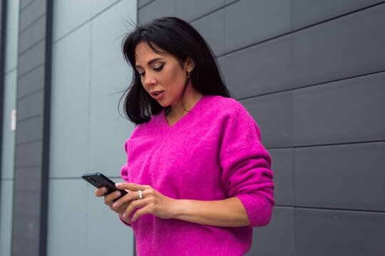 Beautiful woman wearing bright pink sweater posing on dark gray background at city street with smartphone