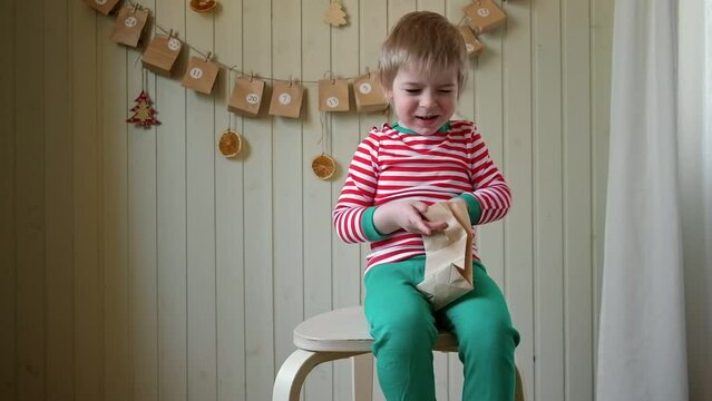 Boy in pajamas, Santa cap eating sweet candy on paper advent calendar with presents background. Little happy child with gift of handmade Christmas calendar on wall. Celebrating at home Xmas tradition. - Powered by Adobe